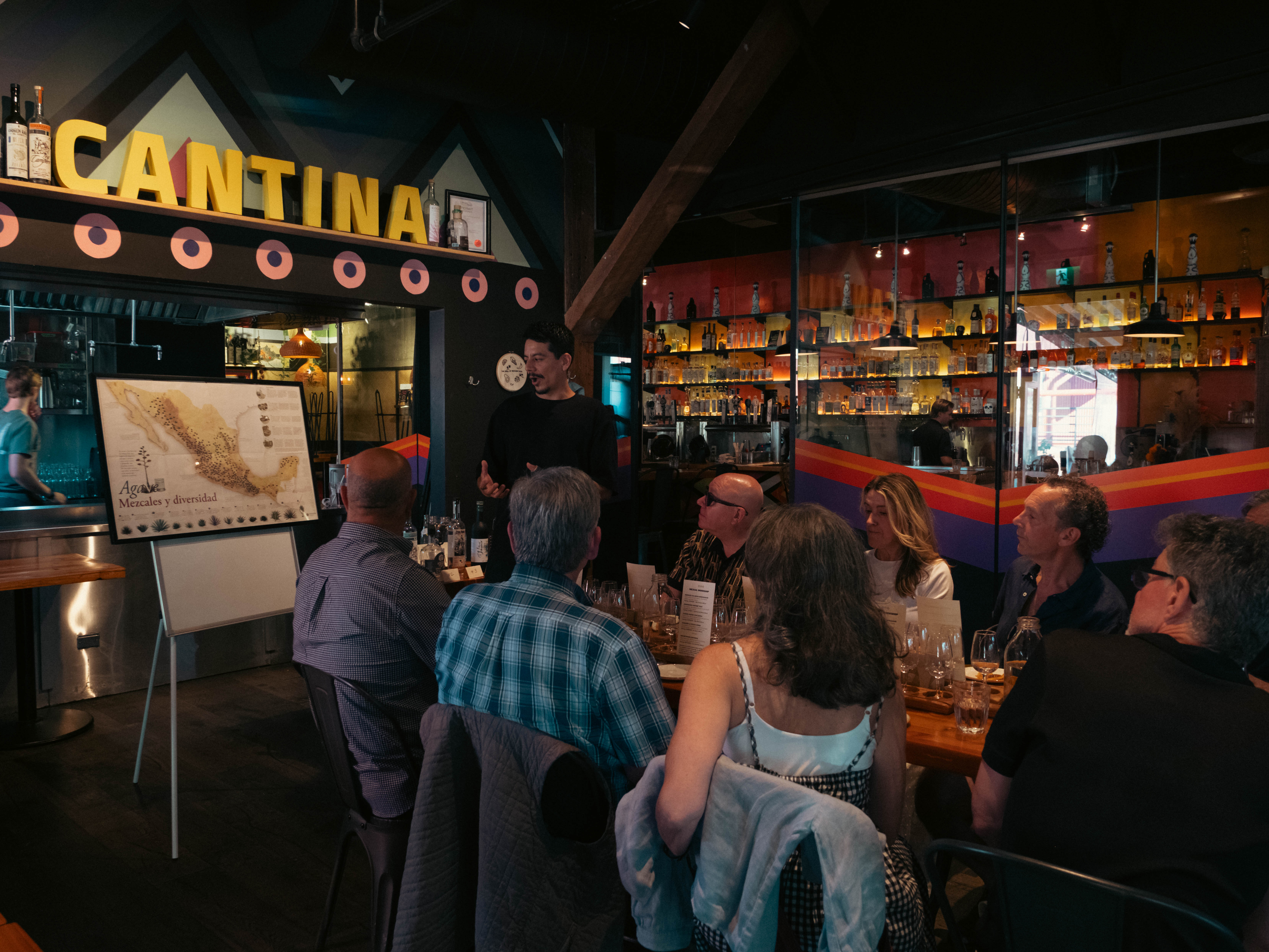 Guests attending an agave workshop inside Alimentaria Mexicana's cantina, with its bottle shelves and CANTINA sign behind them.