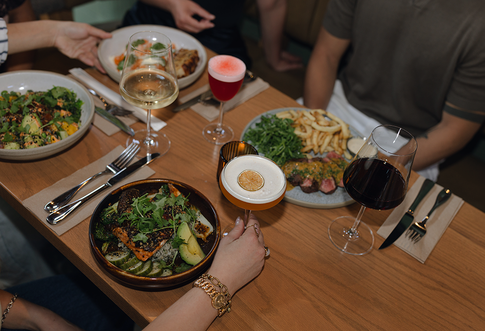 Plates of food and cocktails shared around a table at BREWHALL.