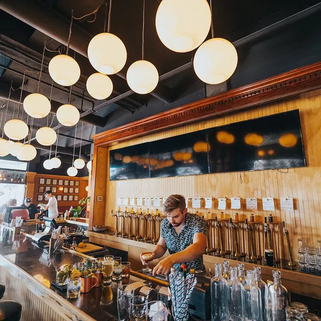 A bartender preparing a drink beneath round pendant lights at Ballyhoo Public House's bar.
