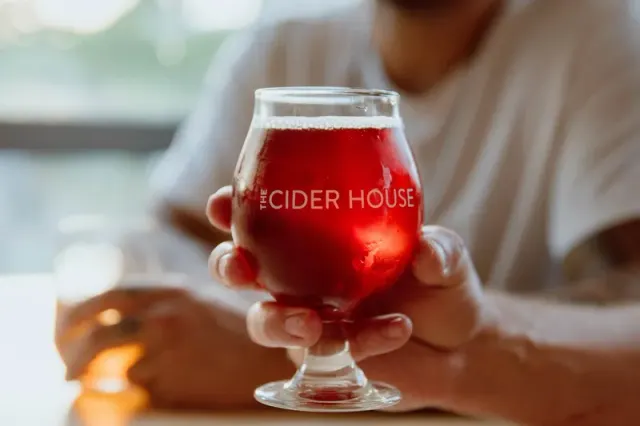 A glass of red cider held above a table at The Cider House.