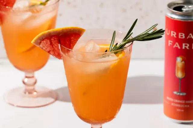 Orange-coloured drinks garnished with citrus and herbs beside a can.