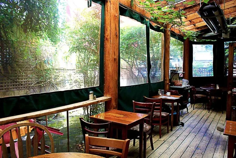 The Naam's covered patio: wooden tables and chairs on a plank floor under a timber trellis, with clear side panels and greenery outside.