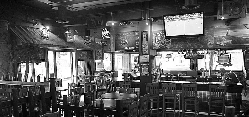 Black and white interior of Two Parrots: wooden tables and bar stools along a tiled bar, televisions overhead and painted signage reading @TwoParrotsVan.